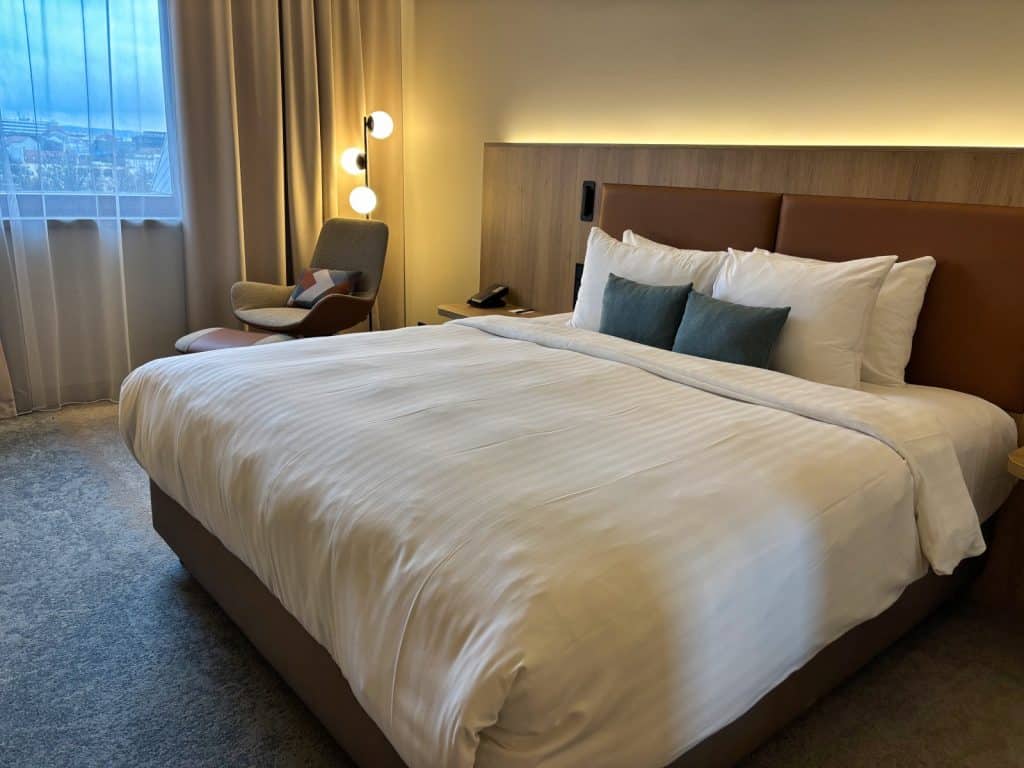 Ein modern eingerichtetes Hotelzimmer im Pilsen Marriott mit einem großen Doppelbett, weißen Laken, dekorativen Kissen und einem gemütlichen Sessel in der Ecke neben einem Fenster mit bodenlangen Vorhängen.