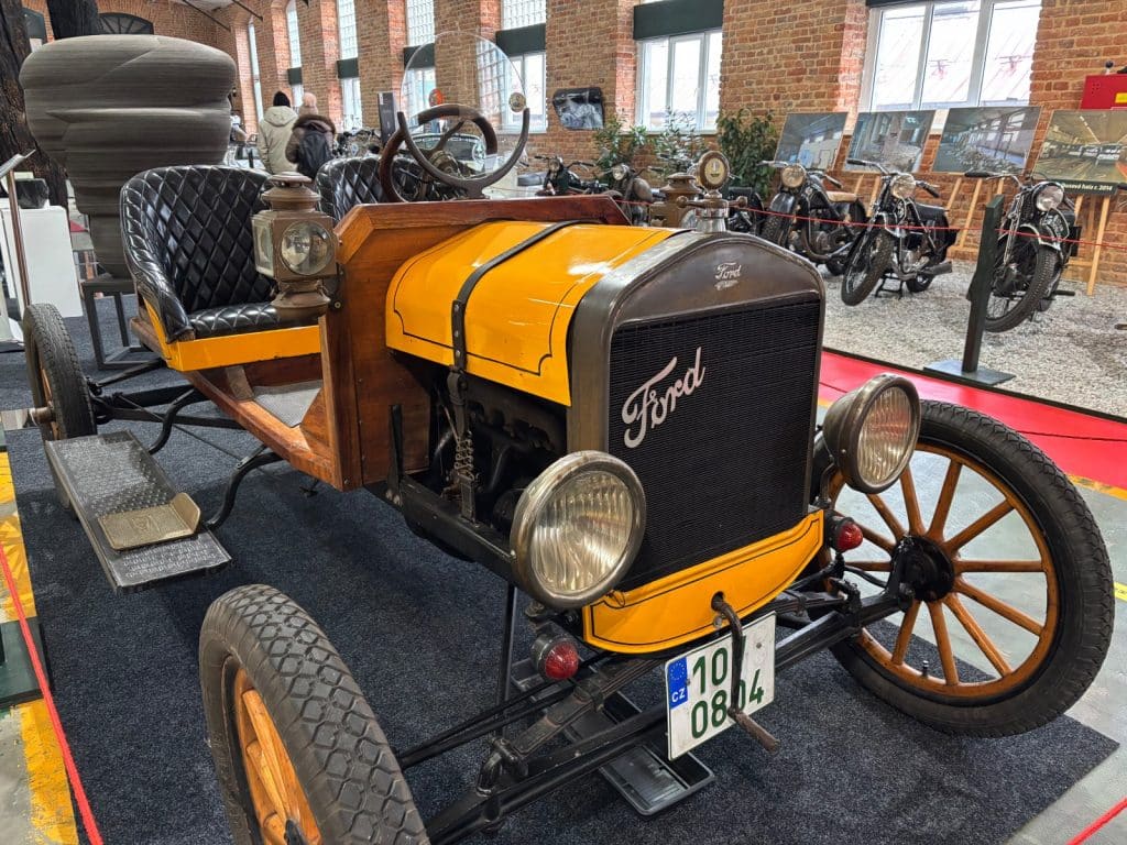 Ein restaurierter, gelber Ford Oldtimer mit Holzkarosserie und schwarzen Ledersitzen steht in der Pilsen Moto Art Ausstellung vor einer Reihe historischer Motorräder.
