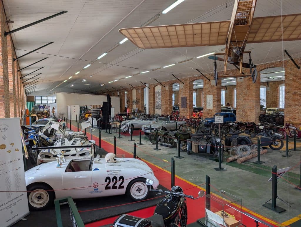 In der Pilsen Moto Art Halle sind zahlreiche historische Fahrzeuge ausgestellt, darunter Oldtimer-Rennwagen, Motorräder mit Beiwagen und ein unter der Decke hängendes Propellerflugzeug.