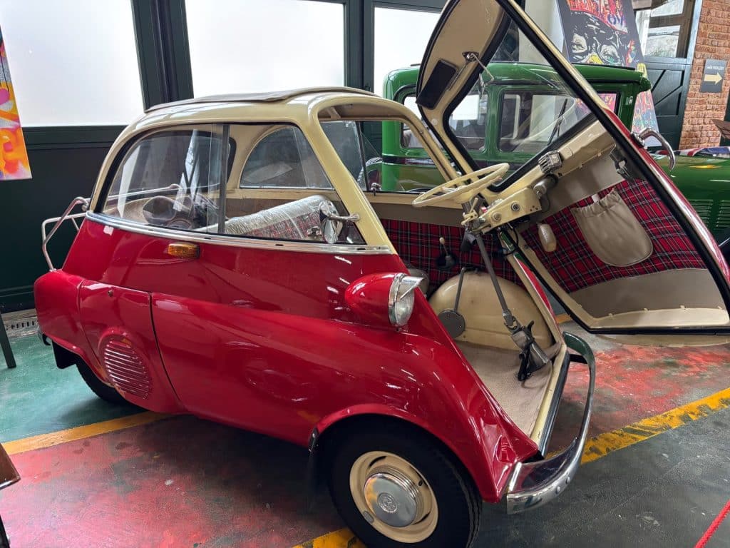 Ein roter BMW Isetta Kleinstwagen mit beigem Dach steht mit weit geöffneter Fronttür in der Pilsen Moto Art Ausstellung und gibt den Blick auf das karierte Interieur und das Lenkrad frei.