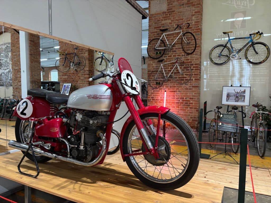 Ein historisches rotes Jawa-Rennmotorrad mit silbernem Tank und der Startnummer 2 steht Moto Art Ausstellung, während im Hintergrund Fahrräder an einer Backsteinwand hängen.