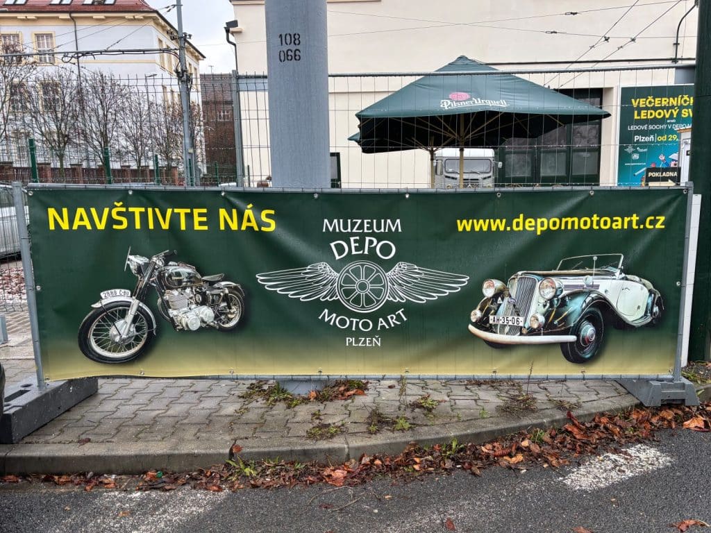 Ein Werbebanner für das Museum „Depo Moto Art“ in Pilsen zeigt ein historisches Motorrad und einen Oldtimer vor grünem Hintergrund mit der Aufschrift „NAVŠTIVTE NÁS“ und der Website-URL.