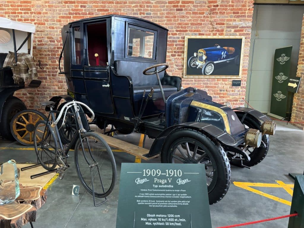 Ein historisches blaues Praga V Automobil aus den Jahren 1909-1910 steht neben einem alten Fahrrad vor einer Ziegelwand. Eine Informationstafel im Vordergrund gibt technische Details zum Fahrzeug an, während im Hintergrund ein gerahmtes Bild eines Rennwagens hängt.