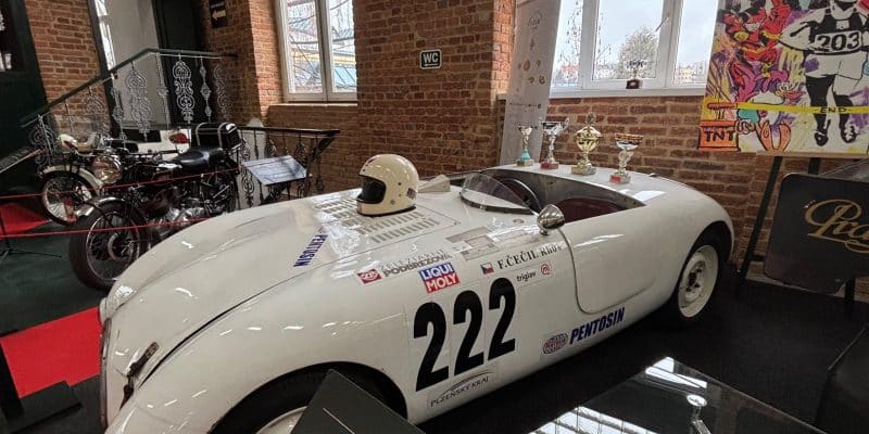 Ein weißer 1950 Hakar Rennwagen mit der Startnummer 222 steht in Pilsen vor einer Backsteinwand, umgeben von Pokalen, einem Helm und historischen Motorrädern.
