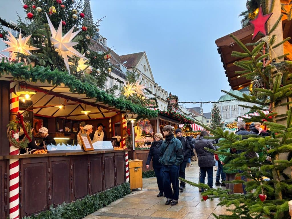 Besucher schlendern an festlich geschmückten Holzbuden mit leuchtenden Herrnhuter Sternen und Tannenreisig auf dem Regensburger Weihnachtsmarkt vorbei. Im Hintergrund ragen historische Gebäude unter einem dämmerigen Abendhimmel auf.