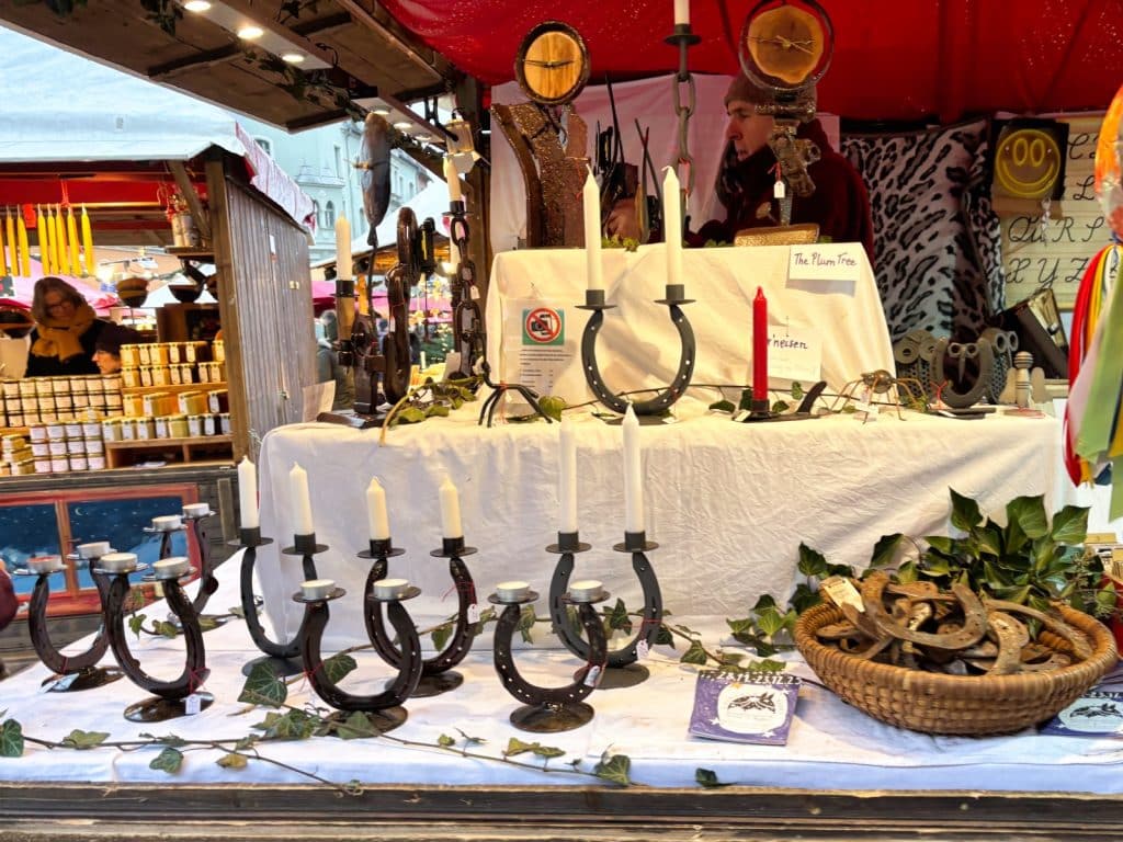 Ein Stand auf dem Regensburger Weihnachtsmarkt präsentiert handgefertigtes Kunsthandwerk, darunter Kerzenständer aus Hufeisen, Metallskulpturen und dekorative Holzarbeiten auf weißen Stofftischen. Im Hintergrund sind ein Kunsthandwerker bei der Arbeit sowie weitere Marktstände mit Honiggläsern und Lichtern zu sehen.