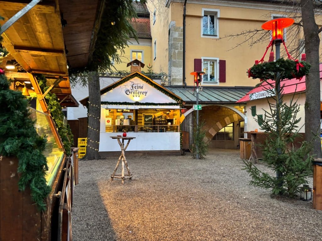 Ein stimmungsvoller Blick auf den Weihnachtsmarkt im Stift in Regensburg mit festlich beleuchteten Holzbuden wie der „Feinen Grillerey“, Stehtischen und weihnachtlicher Dekoration auf einem Kiesplatz vor historischen Gebäuden.