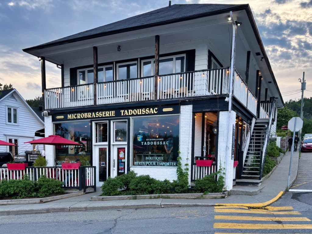 Das zweistöckige weiße Gebäude der Microbrasserie Tadoussac mit schwarzem Dach und Balkon liegt an einer Straßenecke. Vor der Brauerei befindet sich eine kleine Terrasse mit roten Sonnenschirmen und Blumenkästen, während eine Außentreppe in das obere Stockwerk führt.