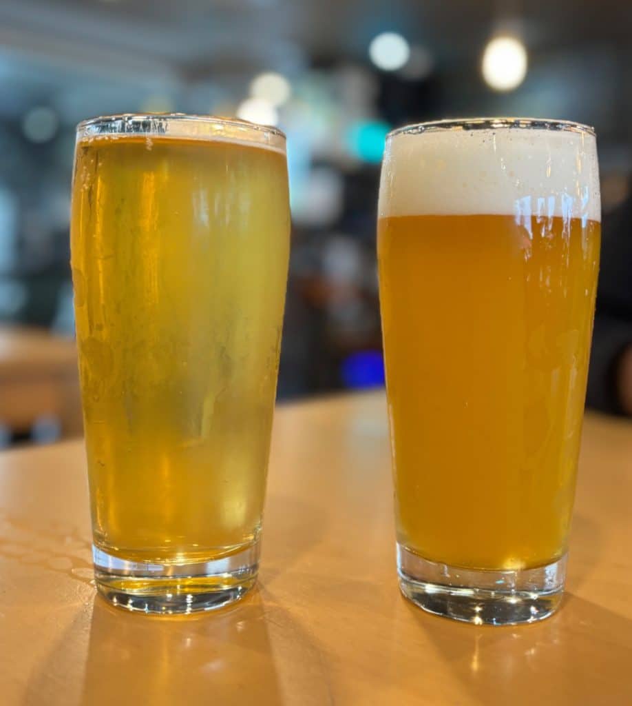 Zwei volle Gläser helles Bier der Microbrasserie Tadoussac stehen nebeneinander auf einem Holztisch, wobei das rechte Glas eine deutlich ausgeprägtere Schaumkrone aufweist.