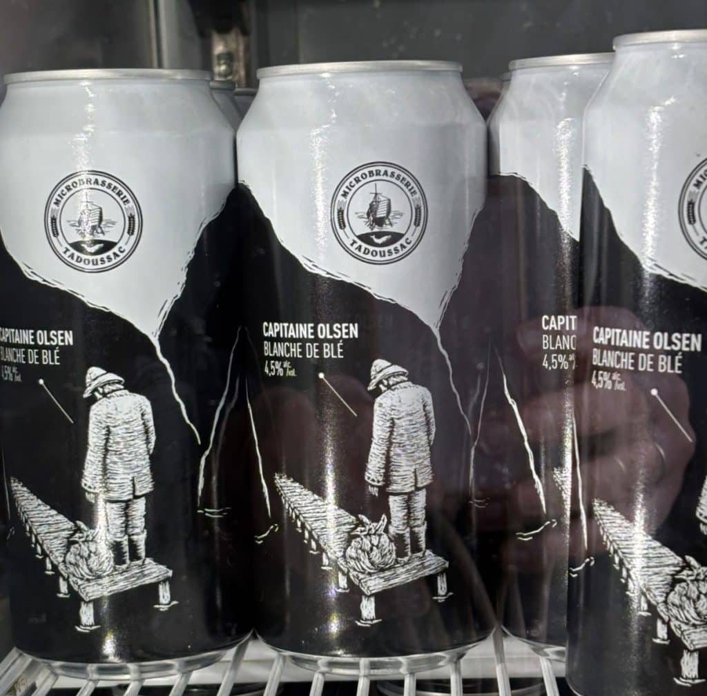 Mehrere Bierdosen der Microbrasserie Tadoussac mit dem Namen „Capitaine Olsen“ zeigen eine schwarz-weiße Illustration eines Seemanns, der auf einem Steg steht und auf das Meer blickt. Das Logo der Brauerei mit einem Segelschiff befindet sich im oberen, weißen Bereich der Dosen.