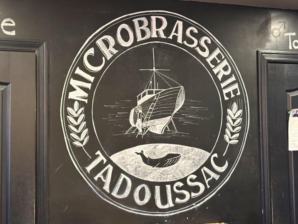 Ein kreisförmiges, mit Kreide gezeichnetes Logo der Microbrasserie Tadoussac auf einer schwarzen Wand zeigt ein Segelschiff, eine Wal-Silhouette und Ähren. Der Schriftzug „MICROBRASSERIE TADOUSSAC“ rahmt die maritimen Motive ein.