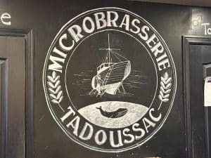 Ein kreisförmiges, mit Kreide gezeichnetes Logo der Microbrasserie Tadoussac auf einer schwarzen Wand zeigt ein Segelschiff, eine Wal-Silhouette und Ähren. Der Schriftzug „MICROBRASSERIE TADOUSSAC“ rahmt die maritimen Motive ein.