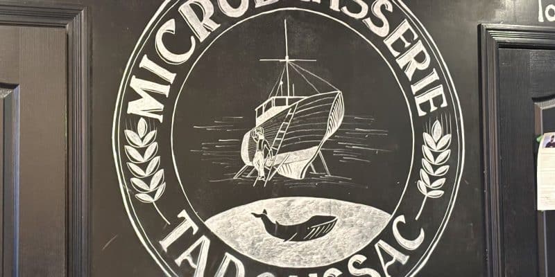 Ein kreisförmiges, mit Kreide gezeichnetes Logo der Microbrasserie Tadoussac auf einer schwarzen Wand zeigt ein Segelschiff, eine Wal-Silhouette und Ähren. Der Schriftzug „MICROBRASSERIE TADOUSSAC“ rahmt die maritimen Motive ein.