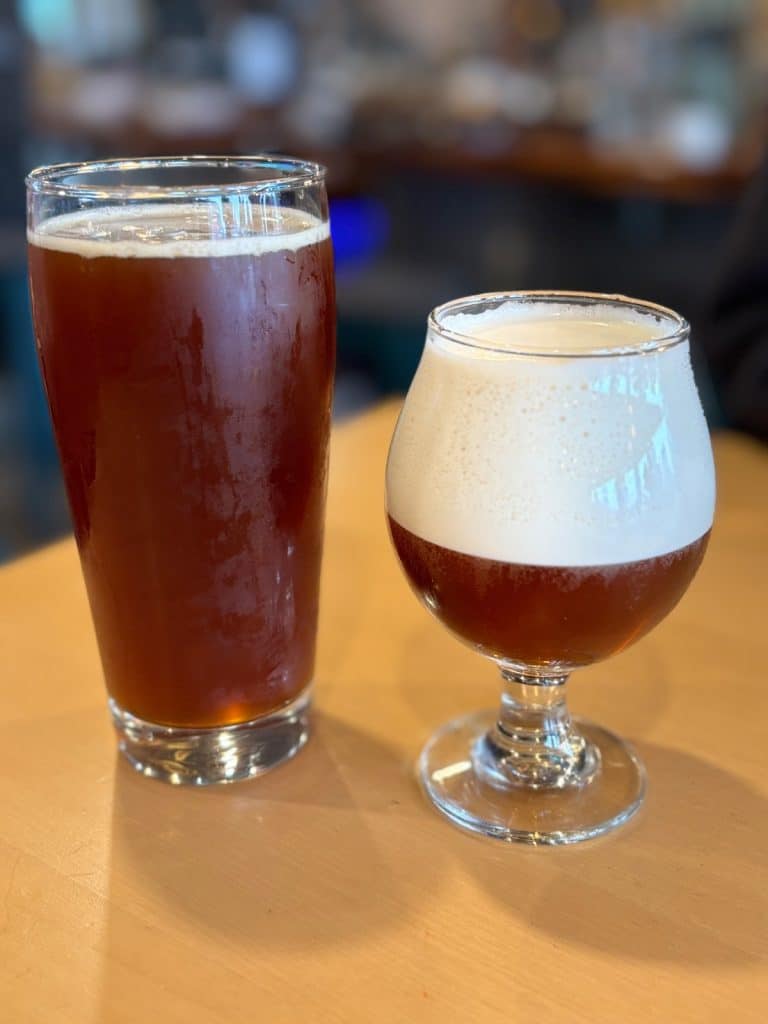 Zwei Gläser mit dunklem Bier der Brauerei in Tadoussac stehen auf einem Holztisch, wobei das linke hohe Glas kaum Schaum und das rechte bauchige Glas eine sehr ausgeprägte, dichte Schaumkrone aufweist.