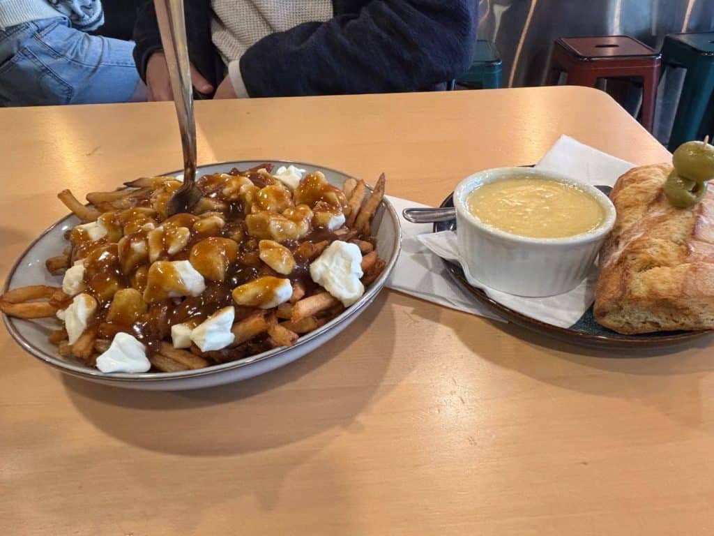 Ein Teller mit Poutine, bestehend aus Pommes frites, Käsebruch und Bratensoße, steht neben einem kleinen Topf Käsefondue mit Baguette und Oliven.
