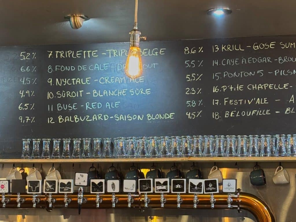 Eine schwarze Kreidetafel in der Microbrasserie Tadoussac listet verschiedene Biersorten mit ihren Namen, Stilen und Alkoholgehalten über einer Reihe von Zapfhähnen und Gläsern auf. Eine hängende Glühbirne beleuchtet die handgeschriebene Menükarte.
