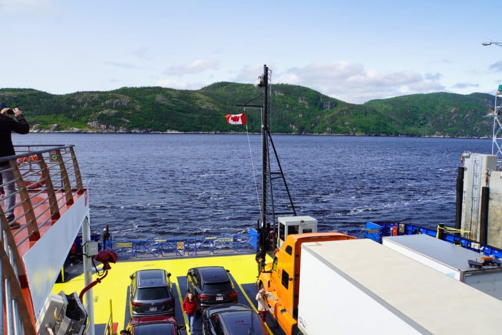 Blick vom Oberdeck der Fähre in Tadoussac auf das Autodeck mit Fahrzeugen und LKWs, während das Schiff vor einer bewaldeten Hügelkette über das Wasser setzt.