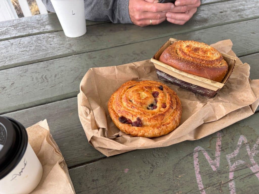 Zwei goldbraune Gebäckstücke, eine Rosinenschnecke und ein süßes Brot in einer Backform, liegen zum Frühstück auf braunem Papier auf einem rustikalen Holztisch neben Kaffeebechern.
