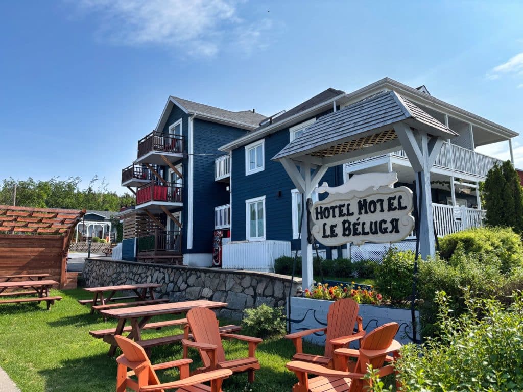 Das Hotel Motel Le Béluga in Tadoussac präsentiert sich als blaues, mehrstöckiges Gebäude mit weißen Balkonen, davor laden orangefarbene Stühle und Picknicktische auf einer Rasenfläche zum Verweilen ein.