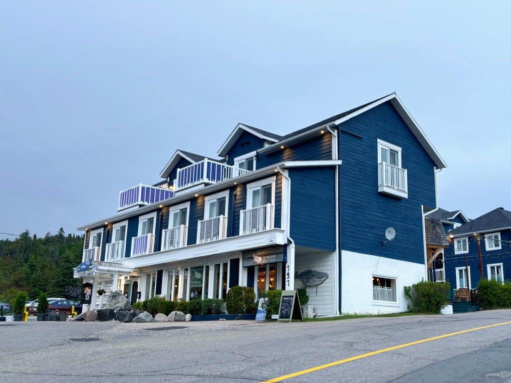 Ein großes, dunkelblaues Gebäude in Tadoussac, das als Hotel und Motel mit Rezeption dient, verfügt über weiße Balkone, eine weiße untere Etage und liegt an einer asphaltierten Straße.
