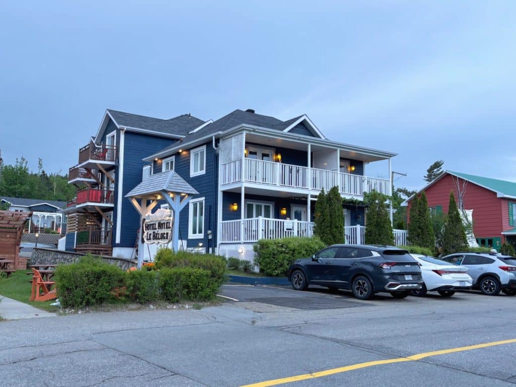 Das Bild zeigt das blaue Gebäude eines Hotels in Tadoussac mit weißen Balkonen, davor geparkten Autos und einem hölzernen Hotelschild im Vordergrund.