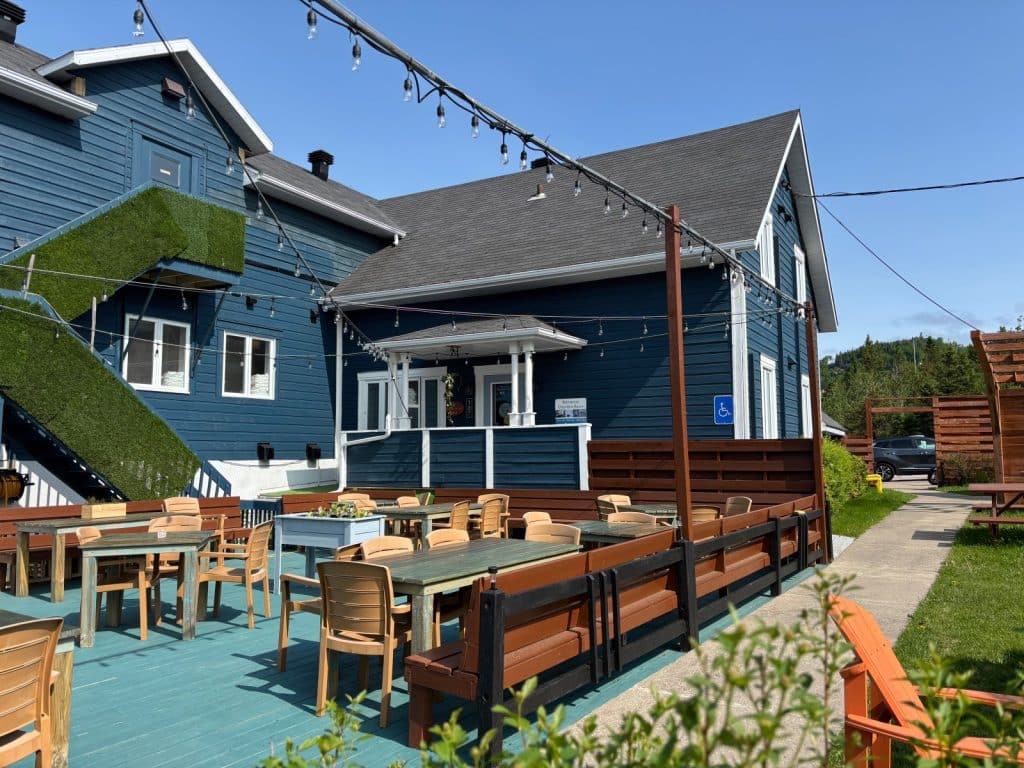 Die Gemeinschaftsterrasse des Motels in Tadoussac ist mit Holztischen, Stühlen und einer Lichterkette ausgestattet und grenzt direkt an das blaue Hauptgebäude mit Außentreppe.