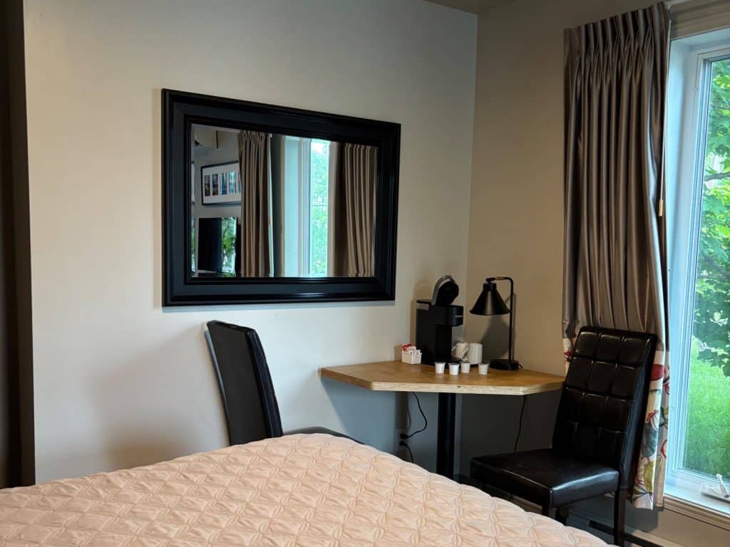 Ein schlicht eingerichtetes Zimmer im Motel mit einem kleinen Holztisch, auf dem eine Kaffeemaschine und Zubehör stehen, flankiert von zwei dunklen Stühlen unter einem großen Wandspiegel.