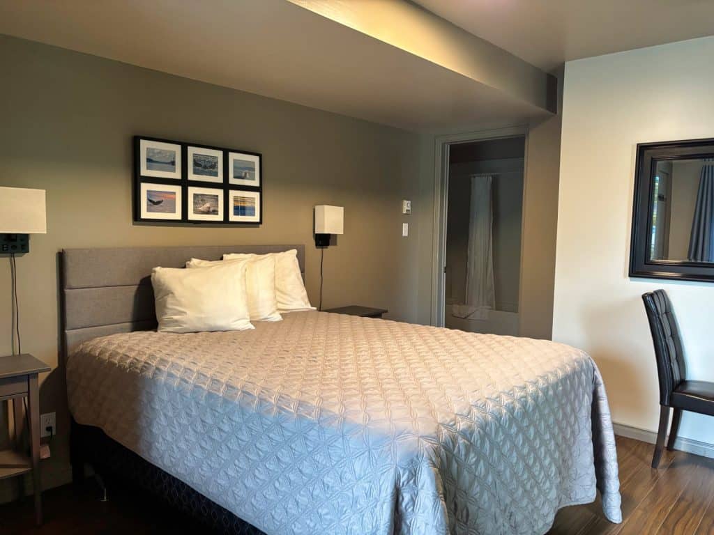 Ein modern eingerichtetes Zimmer im Tadoussac Hôtel-Motel Le Béluga mit einem großen Doppelbett, grauer Steppdecke, gerahmten Naturfotografien an der Wand und Blick in das angrenzende Badezimmer.