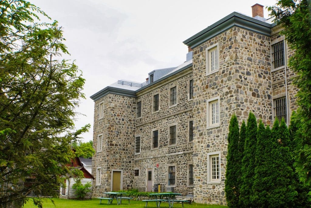 Sehenswürdigkeiten in Trois-Rivières: Das alte Gefängnis von Trois-Rivières ist ein mehrstöckiges, historisches Gebäude aus grauem Naturstein mit vergitterten Fenstern, das von grünen Bäumen und Picknicktischen auf einer Rasenfläche umgeben ist.