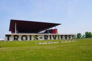 Das Amphithéâtre Cogeco in Trois-Rivières mit seinem markanten, flachen Dach und dem großen, schwarzen Schriftzug „TROIS-RIVIÈRES“ auf einer Betonmauer hinter einer grünen Wiese.