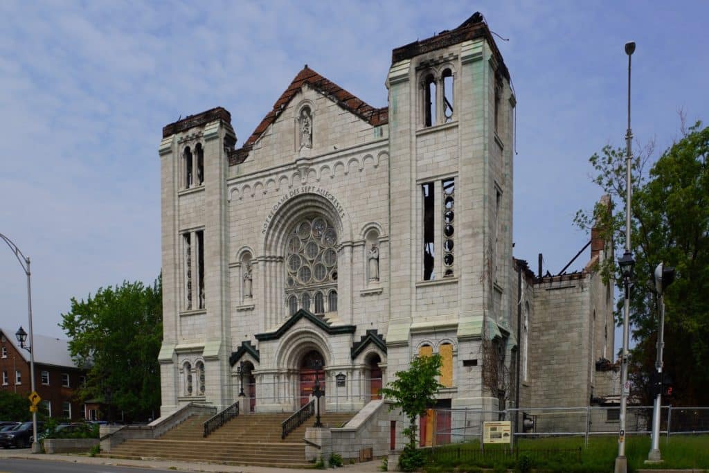 Die Fassade der ausgebrannten Kirche Notre-Dame-des-Sept-Allégresses in Trois-Rivières zeigt schwere Brandschäden, fehlende Dächer an den Türmen und vernagelte Fenster hinter einem Sicherheitszaun. Das steinerne Gebäude weist eine große Fensterrose über dem Haupteingang und leere Glockenstühle in den beschädigten Türmen auf.