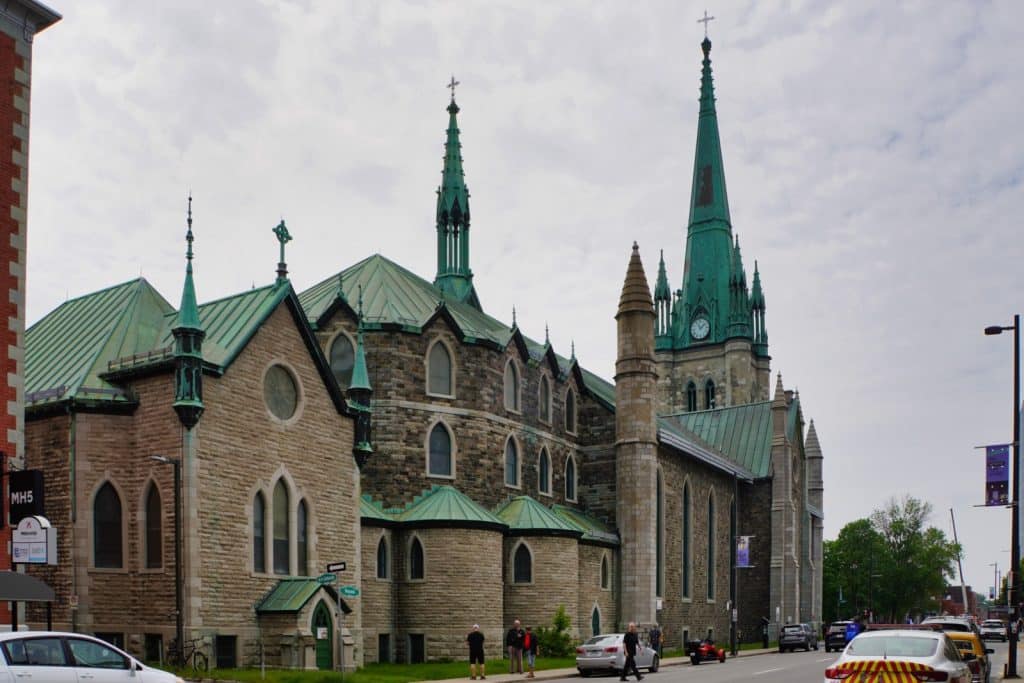 Sehenswürdigkeiten in Trois-Rivières: Die Cathédrale de l'Assomption in Trois-Rivières zeigt sich als imposantes neugotisches Steingebäude mit markanten grünen Kupferdächern, spitzen Türmen und einer Kirchturmuhr unter einem bewölkten Himmel.