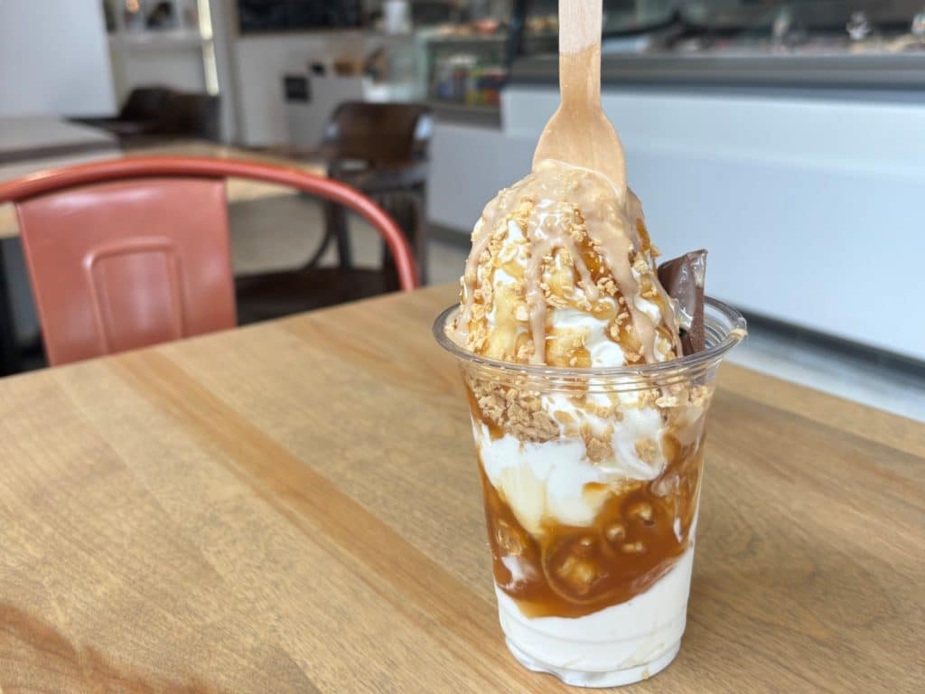 Ein Becher Trois Rivières Eis mit Vanillesoftice, Karamellsauce, Krokant-Streuseln und einem Stück Schokolade.