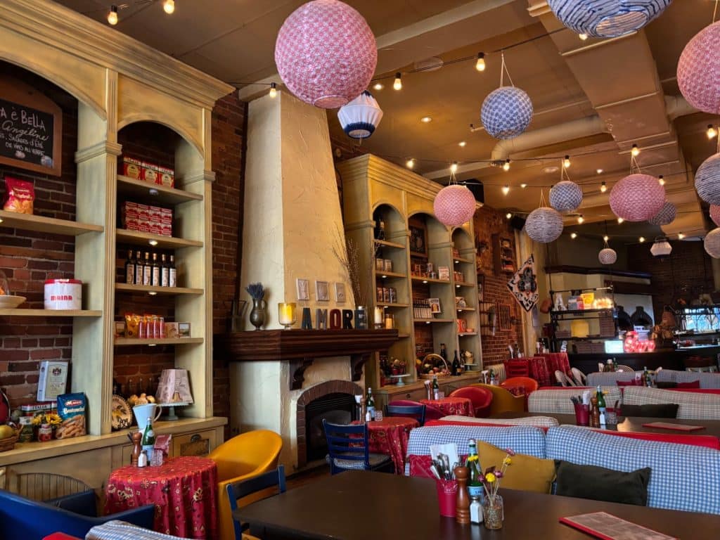 Der gemütliche Innenraum eines italienischen Restaurants in Trois-Rivières ist mit zahlreichen bunten Papierlampions, rustikalen Holzregalen voller Feinkostprodukte und einem Kamin mit der Aufschrift „AMORE“ dekoriert. Die Einrichtung umfasst rot gemusterte Tischdecken, blau-weiß karierte Sitzbänke und eine warme Beleuchtung durch Lichterketten.