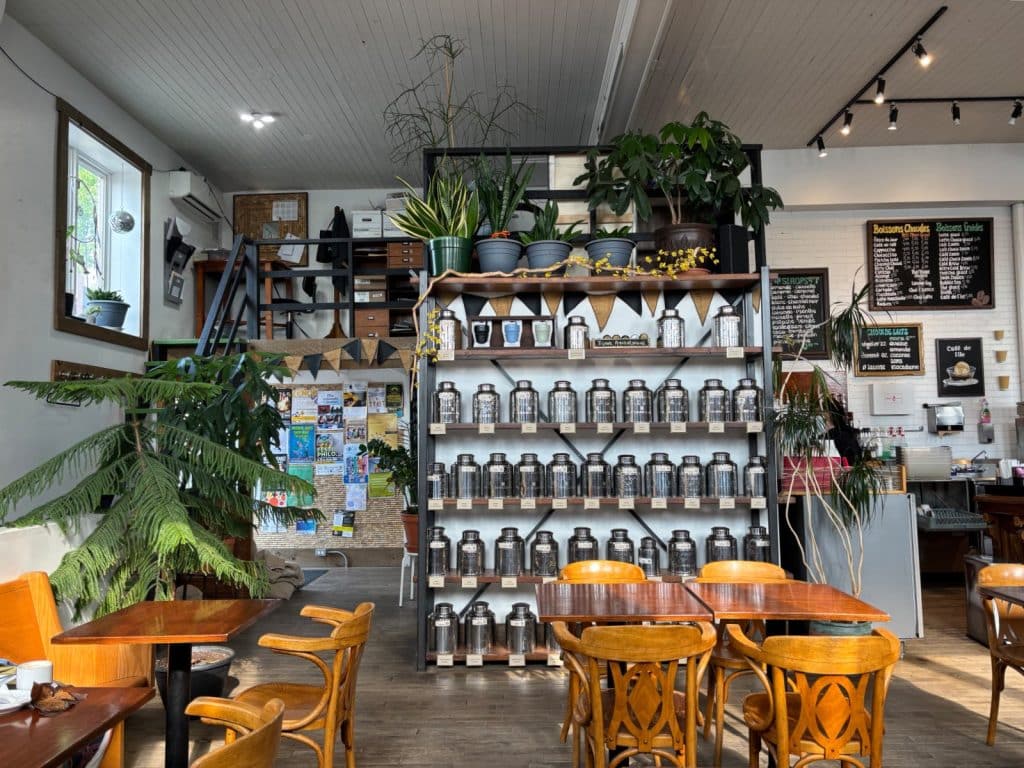 Der gemütliche Innenraum des Cafés Le Caféier in Trois-Rivières zeigt Holztische und -stühle vor einem großen Regal voller Teedosen, ergänzt durch zahlreiche Zimmerpflanzen und eine kleine hölzerne Empore im Hintergrund.