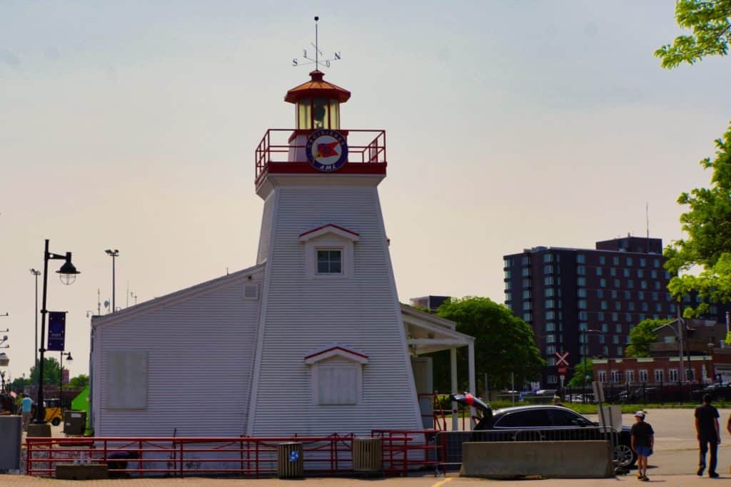 Der weiße, hölzerne Trois-Rivières Leuchtturm steht an einem sonnigen Tag vor einem modernen Stadtpanorama mit einem dunklen Hochhaus im Hintergrund.