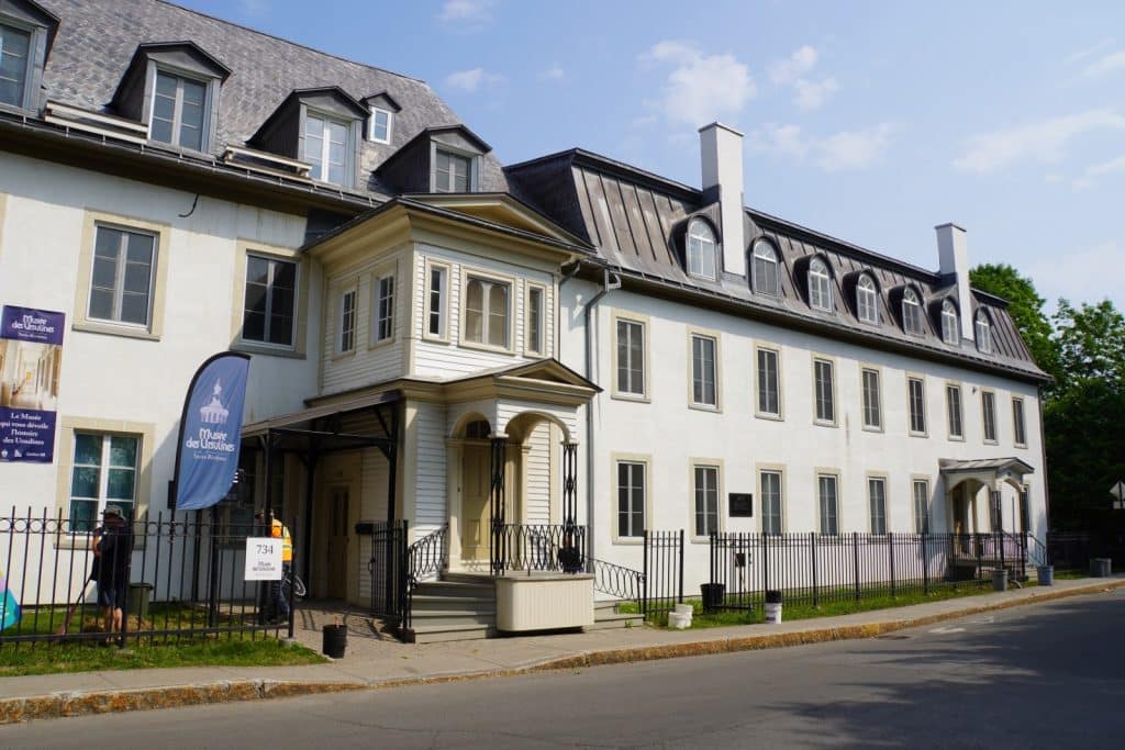 Sehenswürdigkeiten in Trois-Rivières: Das Musée des Ursulines in Trois-Rivières präsentiert sich als historisches, weißes Steingebäude mit einem markanten Mansarddach, zahlreichen Gaubenfenstern und einem hellen hölzernen Eingangsvorbau hinter einem schwarzen Metallzaun.
