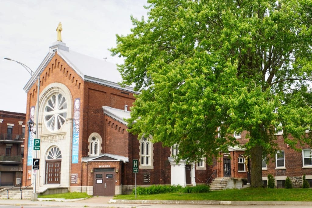 Sehenswürdigkeiten in Trois-Rivières: Das Musée du Père Frédéric Janssoone in Trois-Rivières ist ein rotes Backsteingebäude mit einer markanten Fensterrose und einer goldenen Statue auf dem Dachgiebel, das von einem großen grünen Baum flankiert wird.