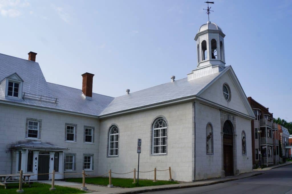 Sehenswürdigkeiten in Trois-Rivières: Das historische Steingebäude der Récollets de Trois-Rivières zeigt eine weiße Kapelle mit einem markanten Glockenturm und Wetterhahn sowie ein angrenzendes Wohnhaus unter einem hellen Metalldach.