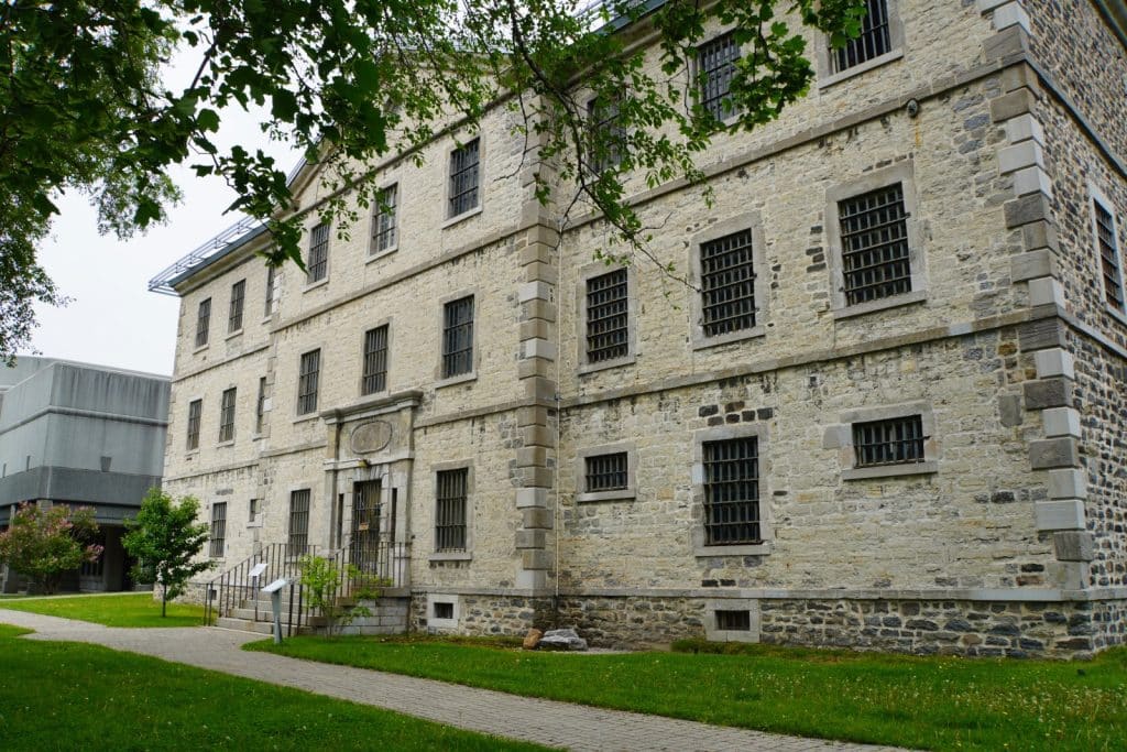 Das historische Steingebäude des alten Gefängnisses von Trois-Rivières zeigt eine massive Fassade mit vergitterten Fenstern und einen zentralen Eingangsbereich mit einer kleinen Treppe. Ein gepflasterter Weg führt an der gepflegten Rasenfläche vor dem monumentalen Bauwerk entlang.