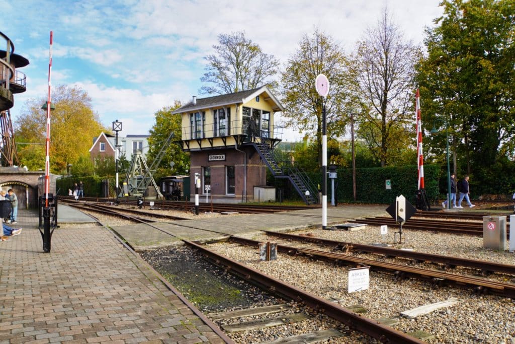 Das Außengelände des Eisenbahnmuseums in Utrecht zeigt ein historisches, zweistöckiges Stellwerk aus Backstein und gelbem Holz, das an mehreren Gleisen mit Weichen, Signalen und Schranken liegt. Im Hintergrund ergänzen herbstliche Bäume und ein alter Kran die museale Bahnanlage unter leicht bewölktem Himmel.