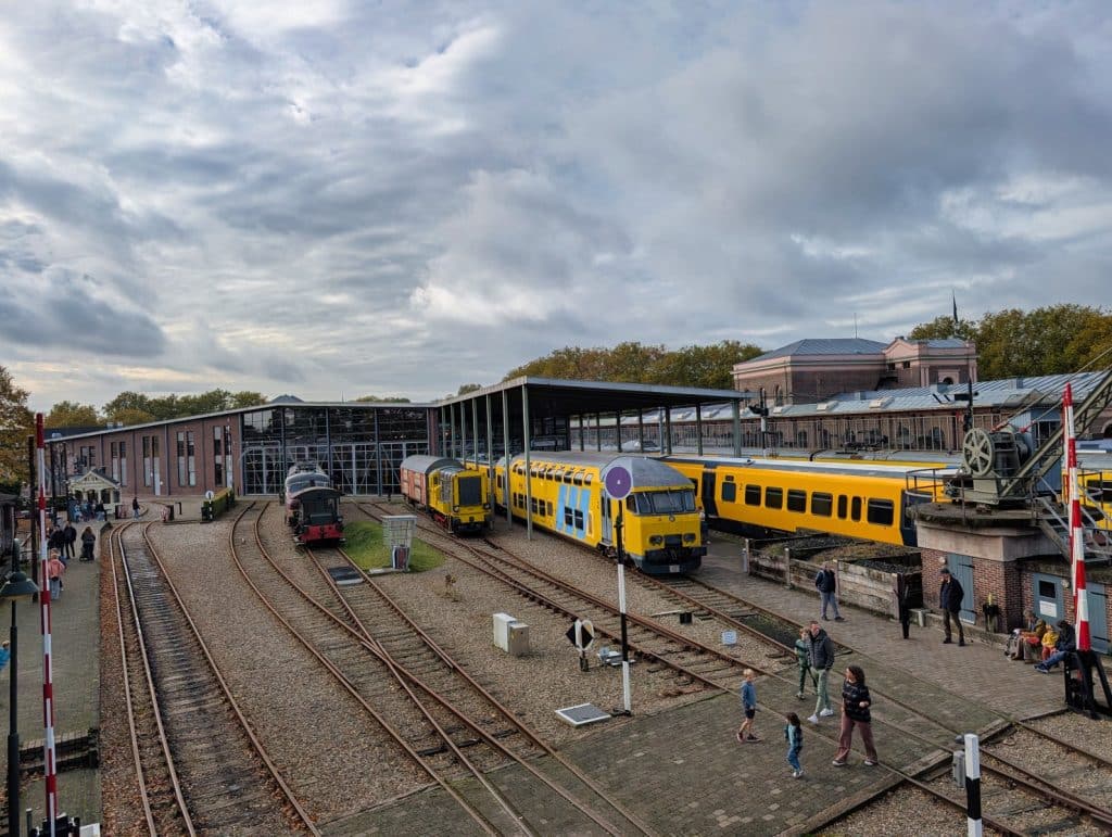 Blick auf das Außengelände des Eisenbahnmuseums in Utrecht mit verschiedenen historischen Zügen auf Gleisanlagen vor einem modernen Glasgebäude. Mehrere Besucher spazieren über das Gelände zwischen den Schienen und Bahnsteigen.