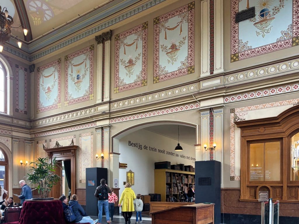Das reich verzierte Innere des historischen Bahnhofsgebäudes in Utrecht zeigt hohe Wände mit kunstvollen Wandmalereien, Rundbögen und wartende Menschen in einer prachtvollen Empfangshalle. Über einem Durchgang prangt eine niederländische Inschrift, während rechts ein hölzerner Fahrkartenschalter das nostalgische Ambiente unterstreicht.