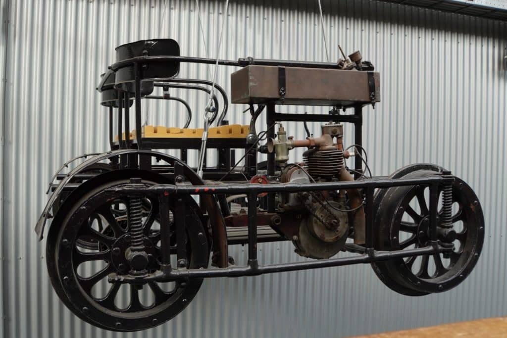Eine historische motorisierte Eisenbahndraisine mit schwarzen Speichenrädern, einem offenen Rohrrahmen und einem sichtbaren Einzylindermotor hängt vor einer silbernen Wellblechwand.