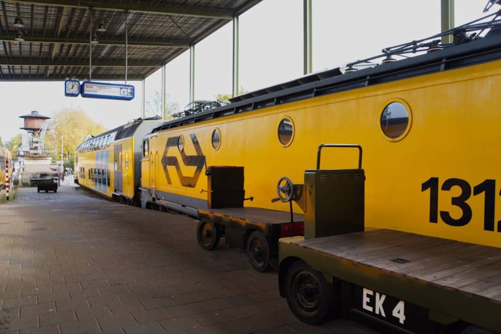 Ein leuchtend gelber historischer Zug der Niederländischen Eisenbahn steht an einem überdachten Bahnsteig in Utrecht, im Vordergrund sind zwei flache Gepäckkarren mit der Aufschrift „EK 4“ zu sehen.