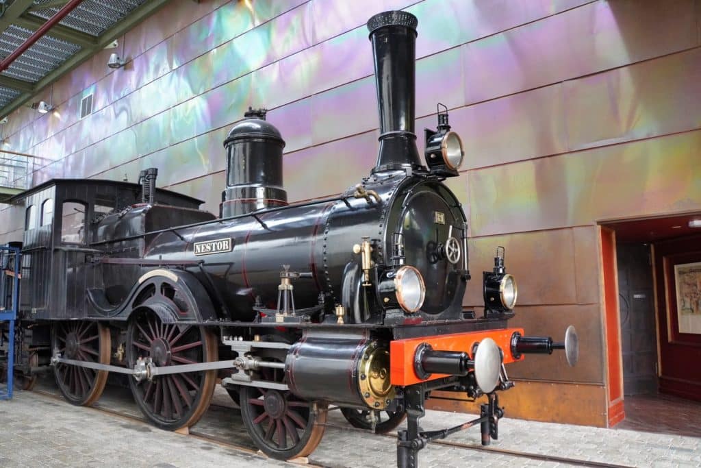 Die historische Dampflokomotive „Nestor“ steht auf Gleisen im Eisenbahnmuseum Utrecht vor einer metallisch schimmernden Wand. Die schwarz glänzende Lokomotive zeichnet sich durch große rote Speichenräder, einen hohen Schornstein und eine markante rote Pufferbohle an der Vorderseite aus.