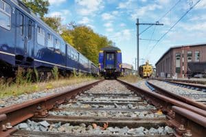 Blick aus einer niedrigen Perspektive auf mehrere Gleise und historische blaue Züge im Eisenbahnmuseum Utrecht unter einem leicht bewölkten Himmel. Rechts im Hintergrund sind ein gelbes Schienenfahrzeug und ein Backsteingebäude zu sehen.