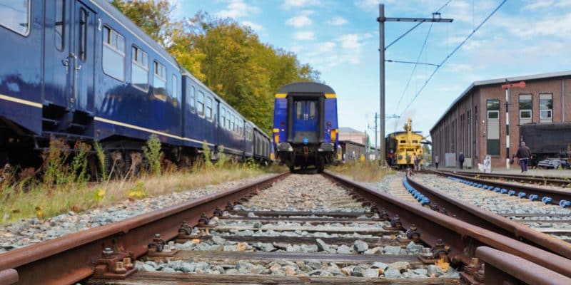 Blick aus einer niedrigen Perspektive auf mehrere Gleise und historische blaue Züge im Eisenbahnmuseum Utrecht unter einem leicht bewölkten Himmel. Rechts im Hintergrund sind ein gelbes Schienenfahrzeug und ein Backsteingebäude zu sehen.