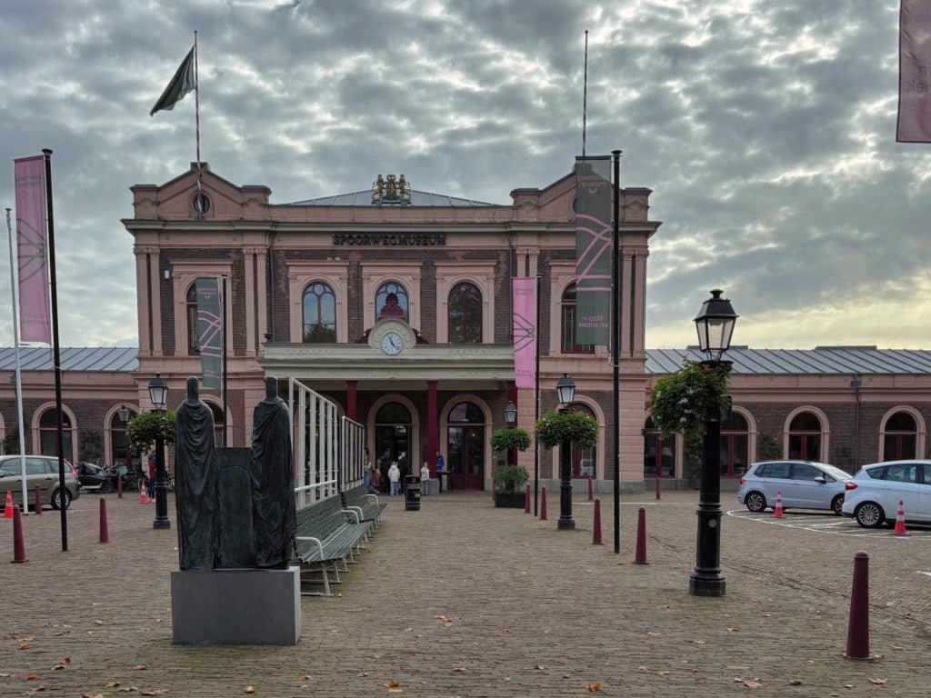 Das historische Empfangsgebäude des Eisenbahnmuseums in Utrecht mit dem Schriftzug „Spoorwegmuseum“ steht hinter einem gepflasterten Vorplatz mit Statuen, Laternen und Fahnen unter einem bewölkten Himmel.