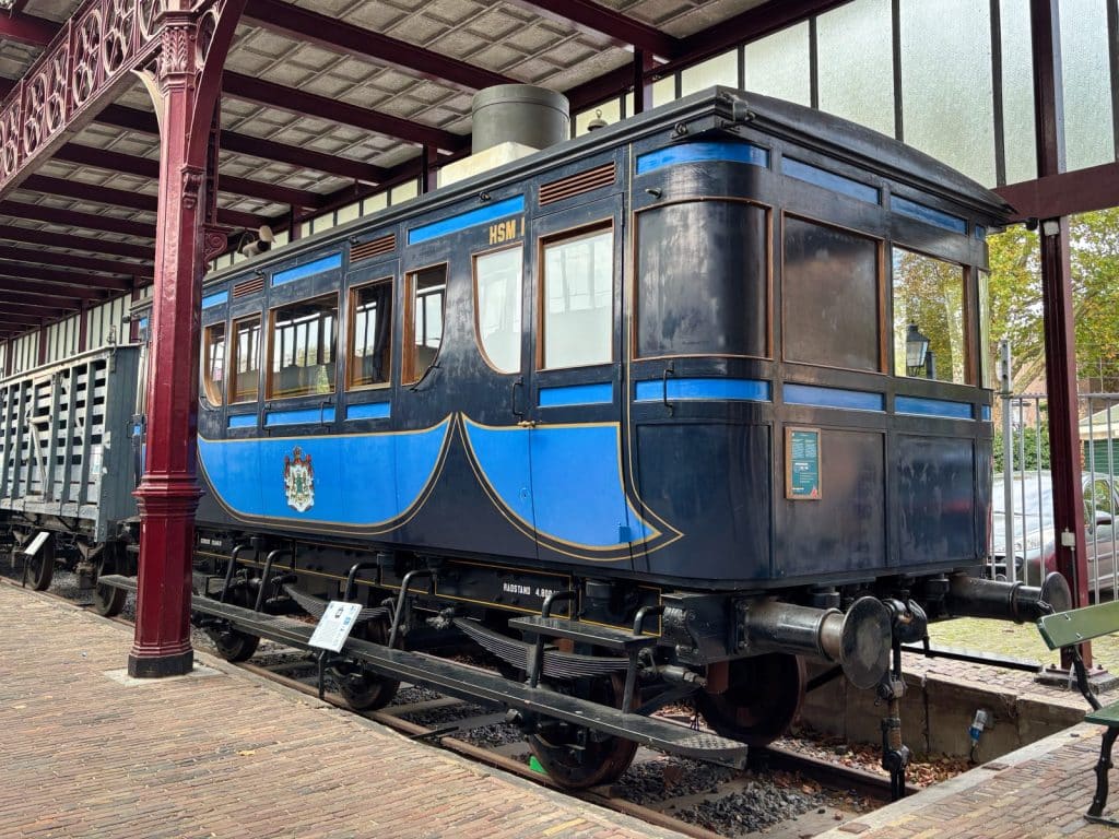 Ein historischer, dunkelblau lackierter Salonwagen der HSM mit hellblauen Verzierungen und dem niederländischen Wappen steht auf Gleisen im Eisenbahnmuseum Utrecht unter einer überdachten Bahnsteighalle.